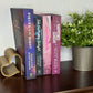 Four romance novels standing upright on a dark wooden shelf, flanked by a gold heart-shaped bookend on the left and a potted green plant on the right. The books feature purple, blue, and pink spines with white and gold typography.