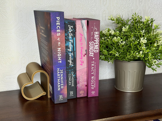Four romance novels standing upright on a dark wooden shelf, flanked by a gold heart-shaped bookend on the left and a potted green plant on the right. The books feature purple, blue, and pink spines with white and gold typography.
