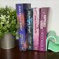 Four romance novels standing upright on a dark wooden table next to a potted green plant and a mint green heart-shaped bookend. 