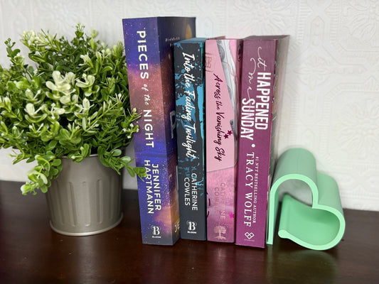 Four romance novels standing upright on a dark wooden table next to a potted green plant and a mint green heart-shaped bookend. 