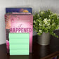 A pink book or journal titled "it HAPPENED" with a stack of mint green sticky notes in front, displayed on a dark wooden shelf next to a potted green plant against a light patterned wall.