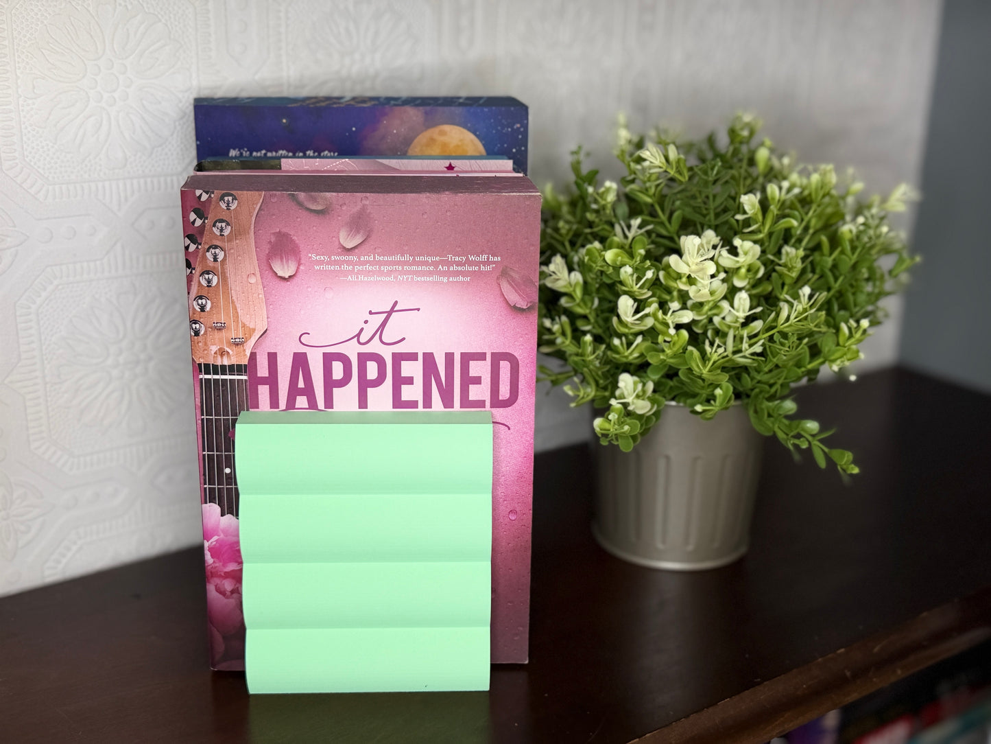 A pink book or journal titled "it HAPPENED" with a stack of mint green sticky notes in front, displayed on a dark wooden shelf next to a potted green plant against a light patterned wall.