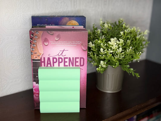 A pink book or journal titled "it HAPPENED" with a stack of mint green sticky notes in front, displayed on a dark wooden shelf next to a potted green plant against a light patterned wall.
