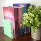 A mint green wavy bookend holding a row of romance novels on a dark wooden table next to a potted green plant.