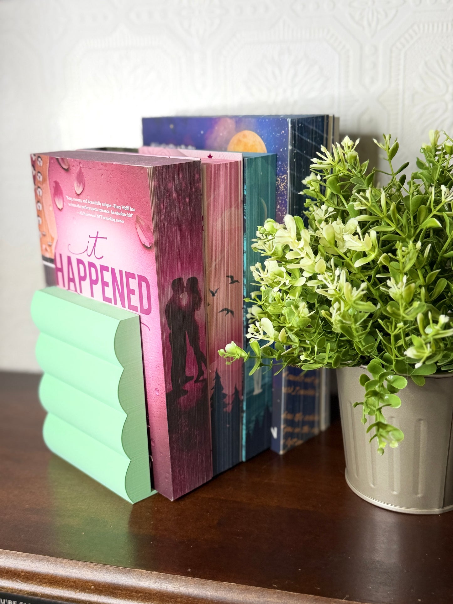 A mint green wavy bookend holding a row of romance novels on a dark wooden table next to a potted green plant.