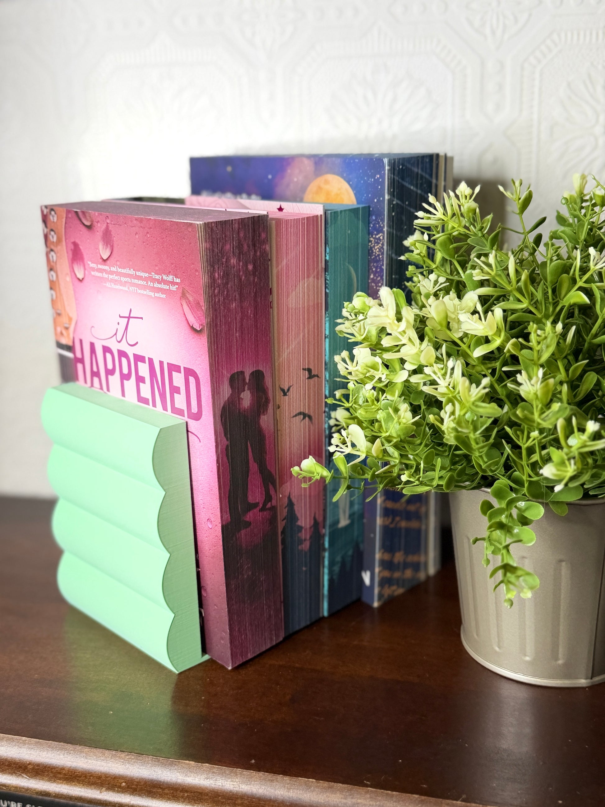 A mint green wavy bookend holding a row of romance novels on a dark wooden table next to a potted green plant.