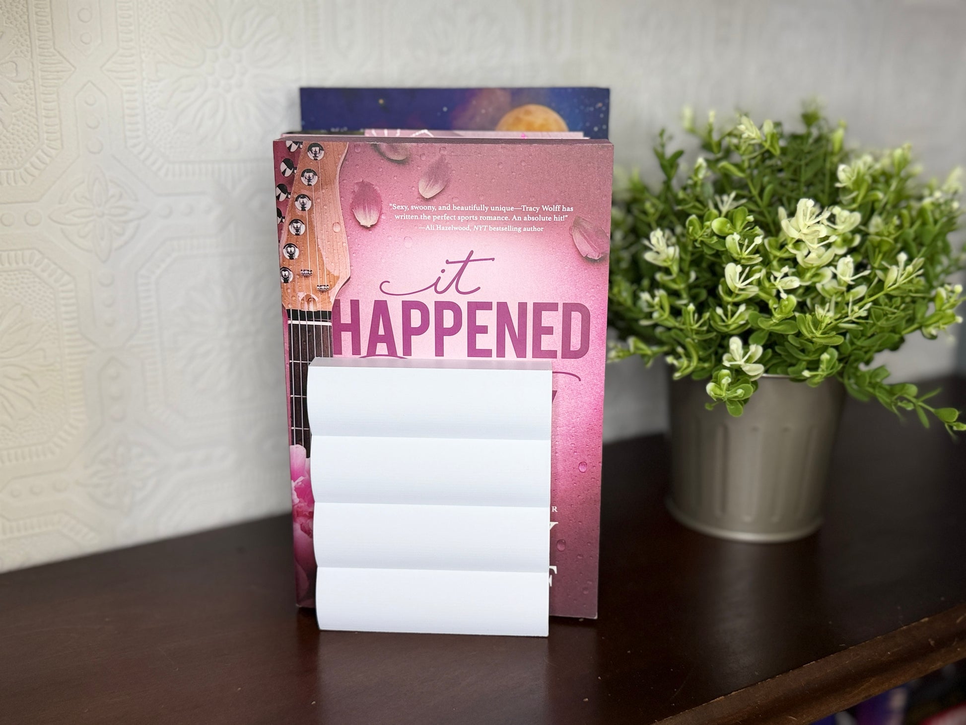 A pink and purple notepad with a guitar and water droplet design on the cover, standing on a dark wooden table next to a potted green plant.