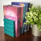 A teal paper bookend shaped like a stack of books holding three romance novels, displayed on a dark wooden table next to a potted green plant.