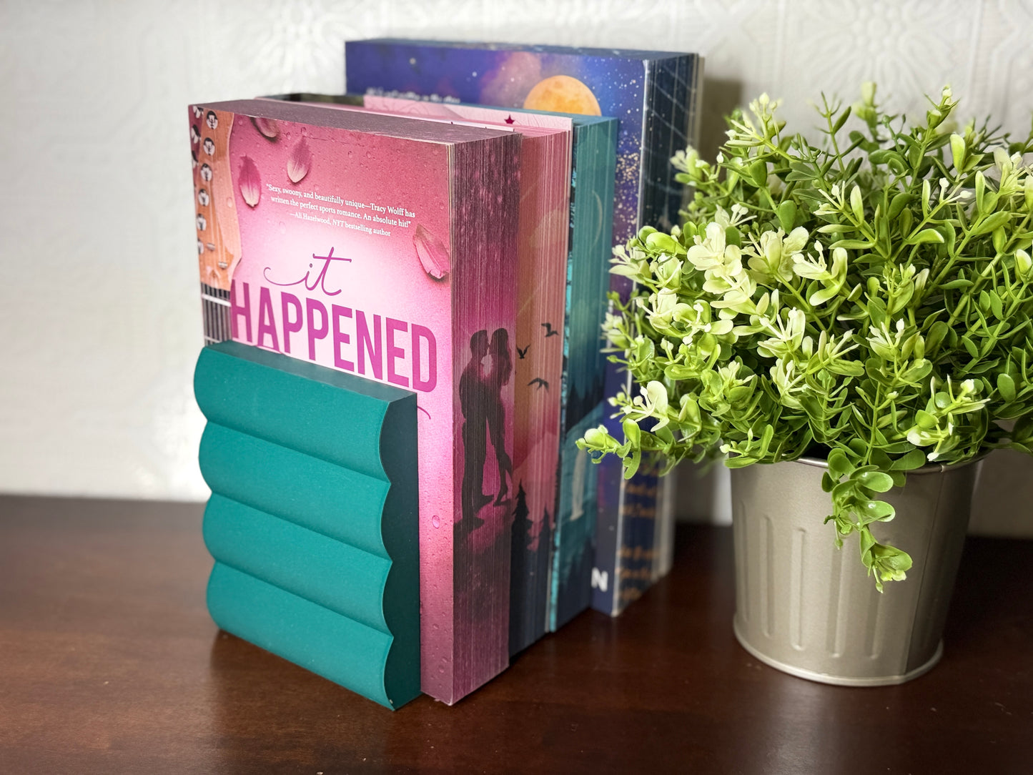 A teal paper bookend shaped like a stack of books holding three romance novels, displayed on a dark wooden table next to a potted green plant.