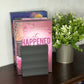 A pink book titled 'it HAPPENED' with a guitar and water droplet design, standing on a dark wooden shelf next to a potted green plant.