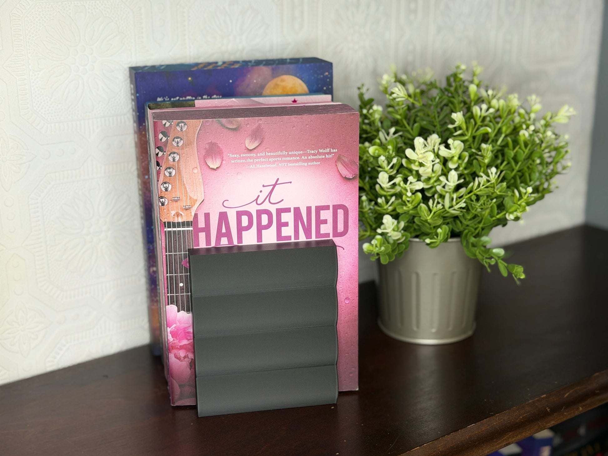A pink book titled 'it HAPPENED' with a guitar and water droplet design, standing on a dark wooden shelf next to a potted green plant.