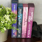 Four books standing upright on a dark wooden shelf next to a potted green plant and a black heart-shaped object, against a light patterned wall.