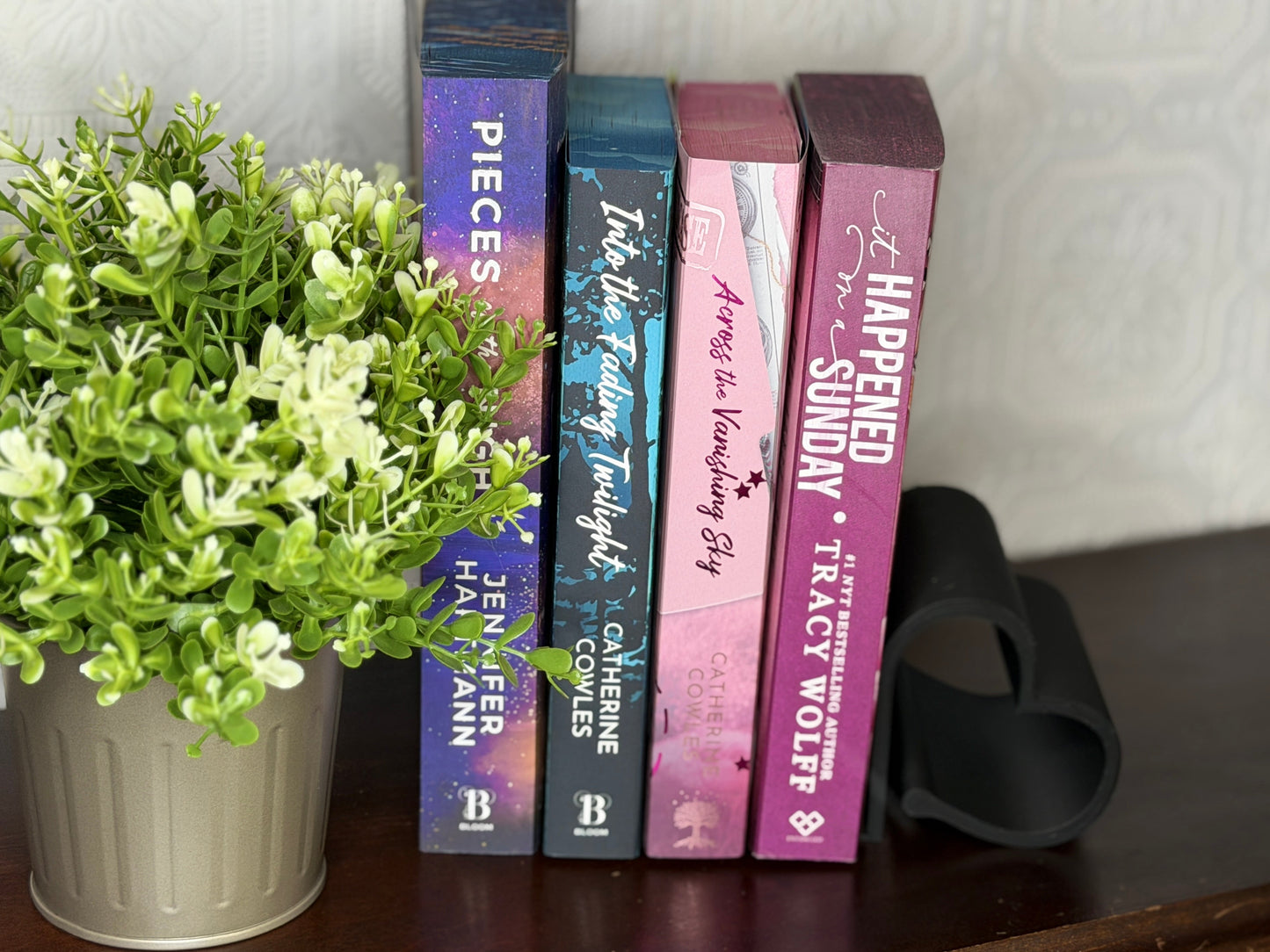 Four books standing upright on a dark wooden shelf next to a potted green plant and a black heart-shaped object, against a light patterned wall.