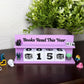 A lavender purple tabletop calendar display with the text 'Books Read This Year' and block numbers showing 0015. The calendar is styled on a dark wooden surface with a potted green plant in the background and small decorative figurines around it.