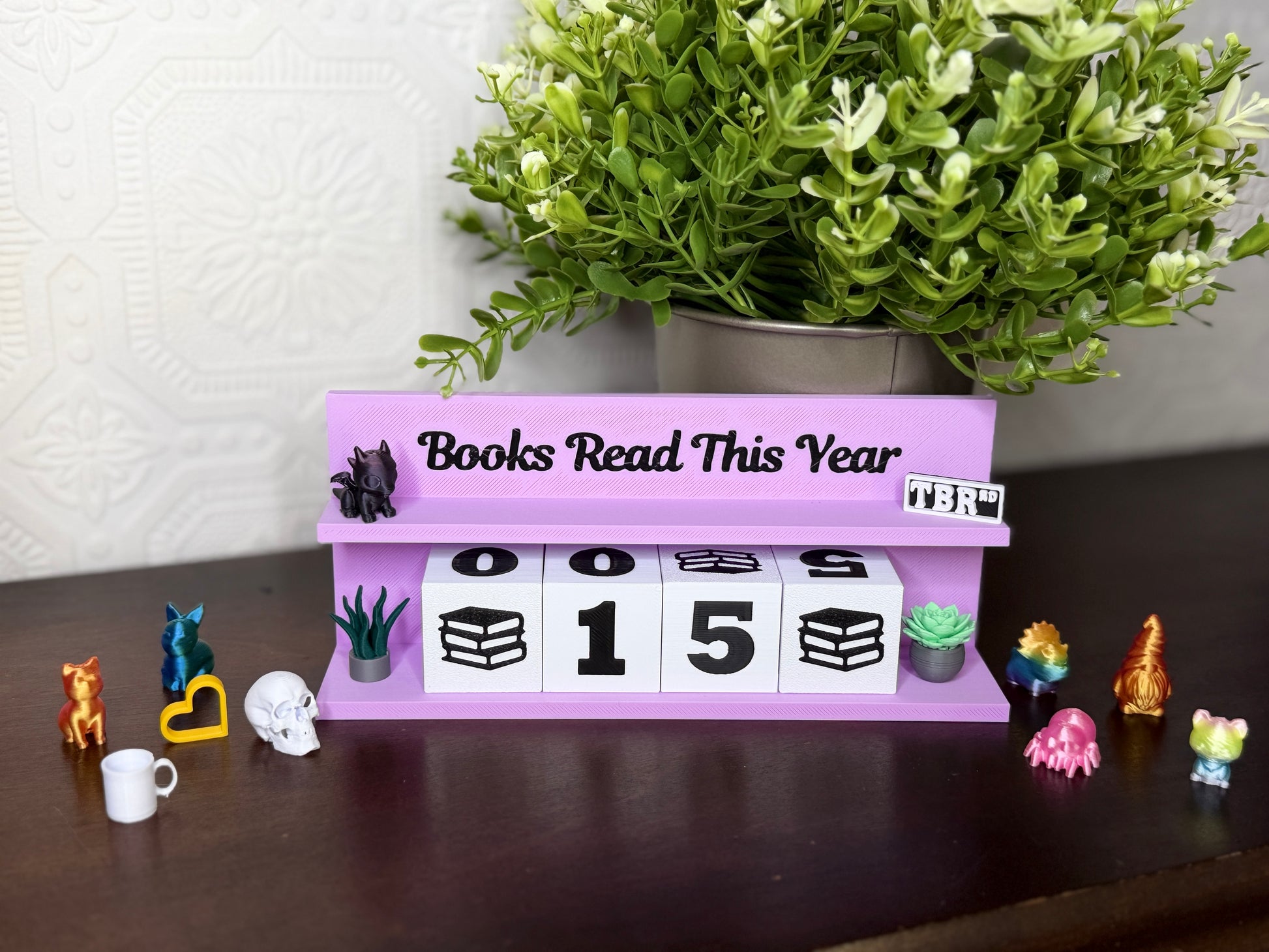 A lavender purple tabletop calendar display with the text 'Books Read This Year' and block numbers showing 0015. The calendar is styled on a dark wooden surface with a potted green plant in the background and small decorative figurines around it.