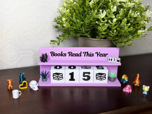 A lavender purple tabletop calendar display with the text 'Books Read This Year' and block numbers showing 0015. The calendar is styled on a dark wooden surface with a potted green plant in the background and small decorative figurines around it.