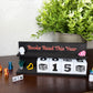 A tabletop display featuring a black wooden sign that reads 'Books Read This Year' in coral lettering, with white flip-down blocks showing '0015' and small decorative figurines around it. A potted green plant sits behind the sign on a light wall, and the setup is placed on a dark wooden surface.