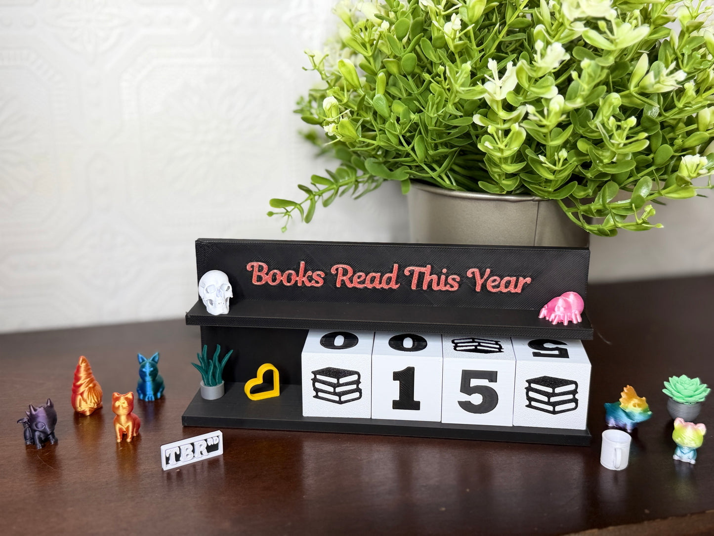 A tabletop display featuring a black wooden sign that reads 'Books Read This Year' in coral lettering, with white flip-down blocks showing '0015' and small decorative figurines around it. A potted green plant sits behind the sign on a light wall, and the setup is placed on a dark wooden surface.