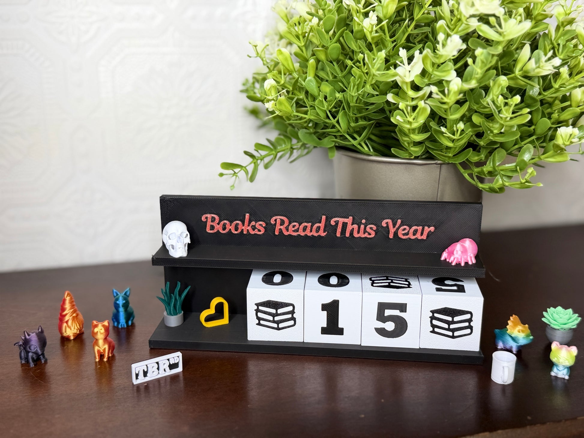 A tabletop display featuring a black wooden sign that reads 'Books Read This Year' in coral lettering, with white flip-down blocks showing '0015' and small decorative figurines around it. A potted green plant sits behind the sign on a light wall, and the setup is placed on a dark wooden surface.