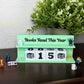 A mint green wooden 'Books Read This Year' calendar stand on a dark brown table, with white cube blocks displaying '00', '15', and '00'. A potted green plant sits behind it against a light patterned wall, with small decorative figurines around the base.