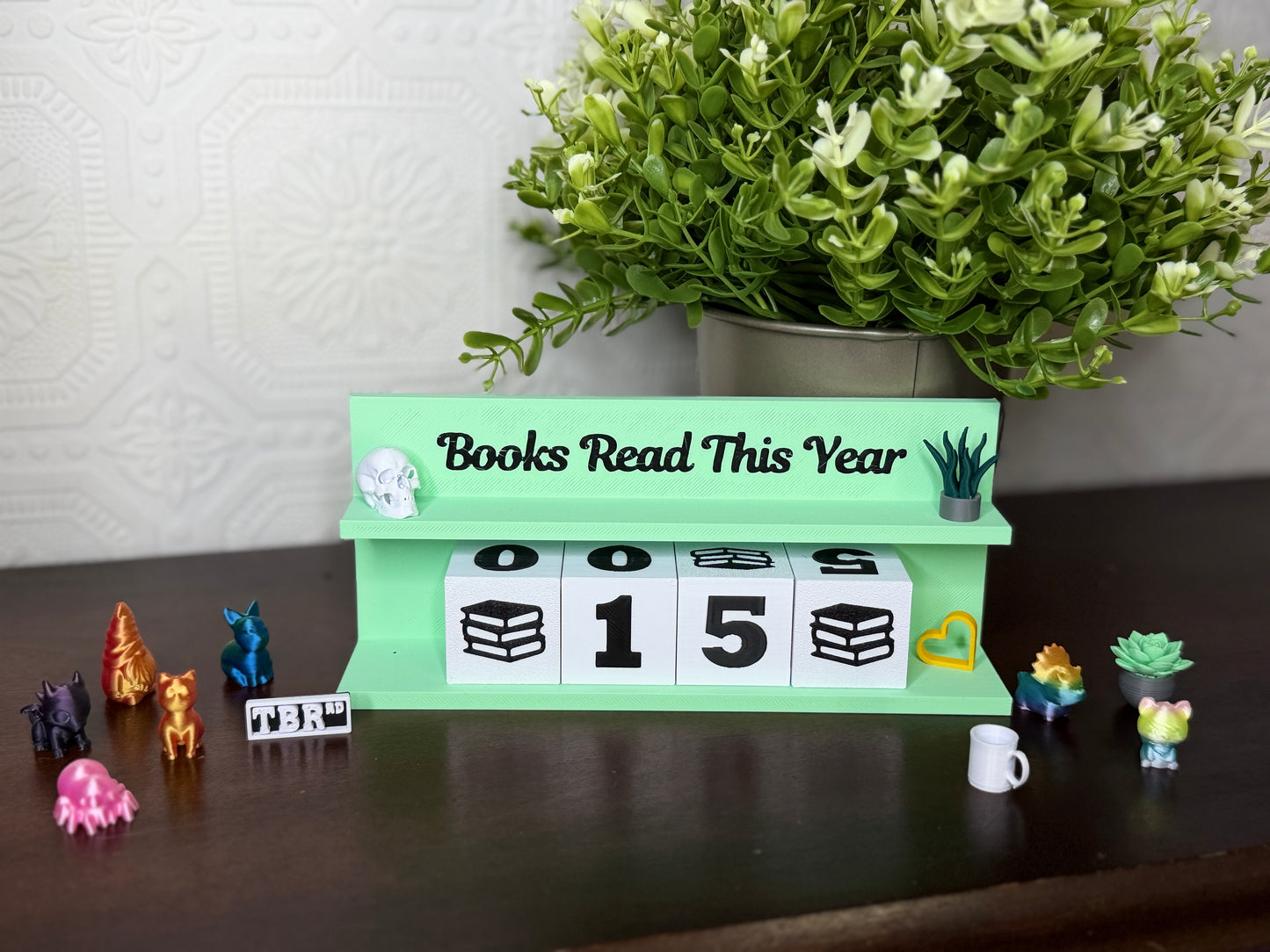A mint green wooden 'Books Read This Year' calendar stand on a dark brown table, with white cube blocks displaying '00', '15', and '00'. A potted green plant sits behind it against a light patterned wall, with small decorative figurines around the base.