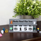 A gray wooden block calendar sign on a dark wooden table, displaying 'Books Read This Year' with the numbers 0, 0, 1, 5. The sign is styled with small decorative figurines and a potted green plant in the background.