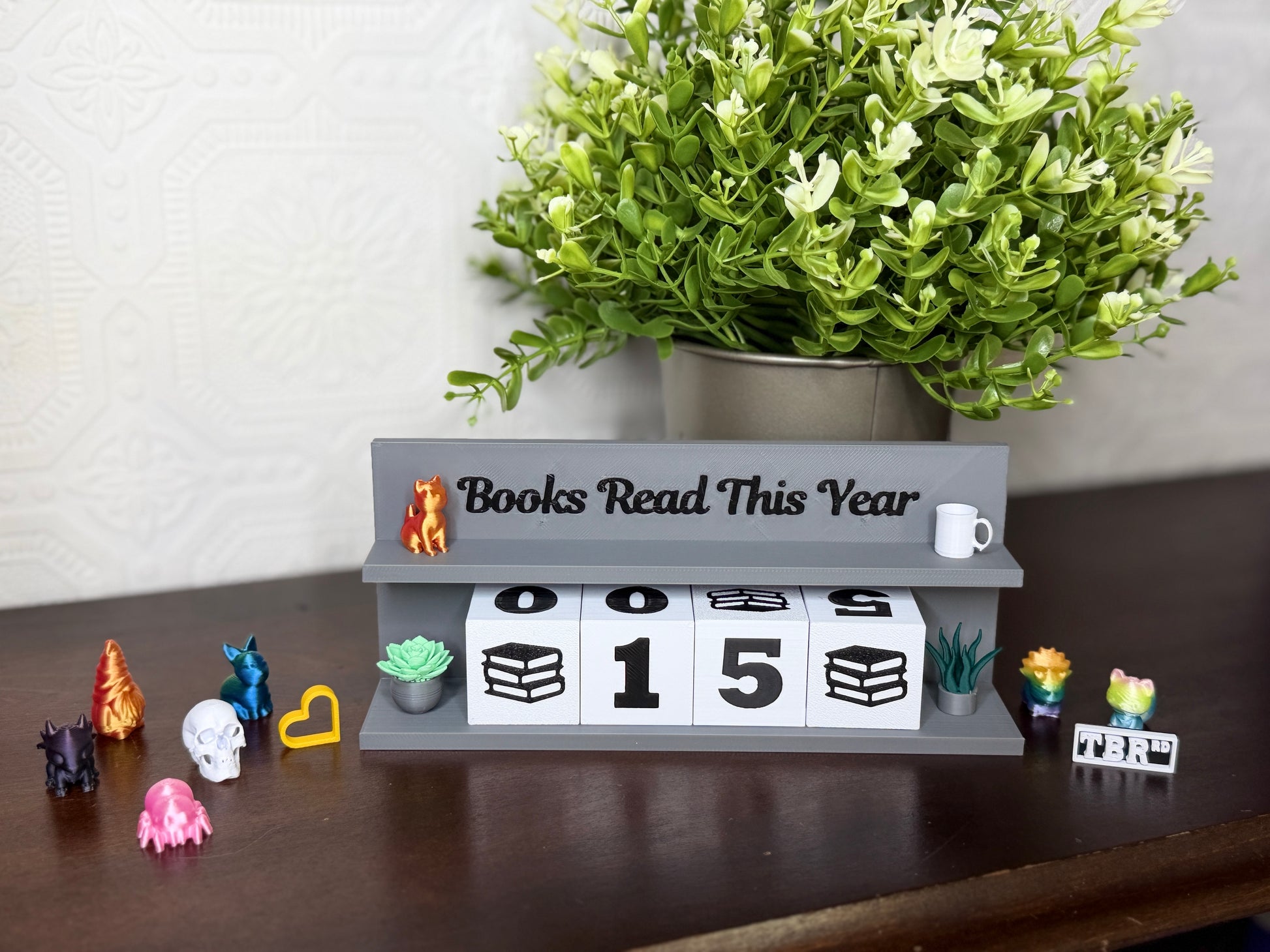 A gray wooden block calendar sign on a dark wooden table, displaying 'Books Read This Year' with the numbers 0, 0, 1, 5. The sign is styled with small decorative figurines and a potted green plant in the background.