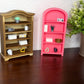 Two miniature dollhouse bookshelves, one gold and one pink, styled with tiny decorative items on a dark wooden table against a textured white wall.
