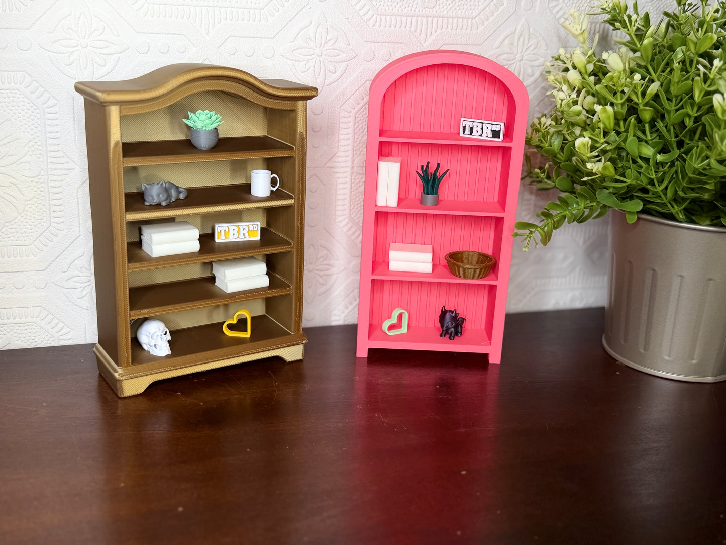 Two miniature dollhouse bookshelves, one gold and one pink, styled with tiny decorative items on a dark wooden table against a textured white wall.