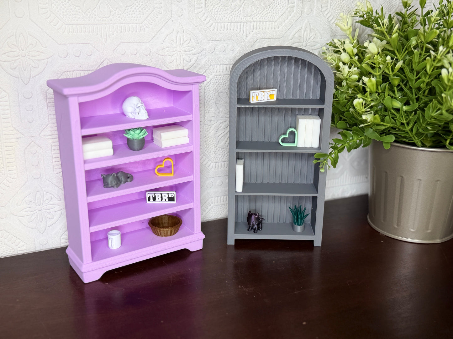 Two miniature toy bookshelves, one lavender purple and one gray, displayed on a dark wooden surface against a white patterned wall, with a potted green plant on the right.