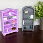 Two miniature toy bookshelves, one lavender purple and one gray, displayed on a dark wooden surface against a white patterned wall, with a potted green plant on the right.