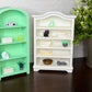 Two miniature decorative bookshelves, one mint green and one white, styled with tiny books, plants, and small figurines on a dark wooden surface against a textured white wall.