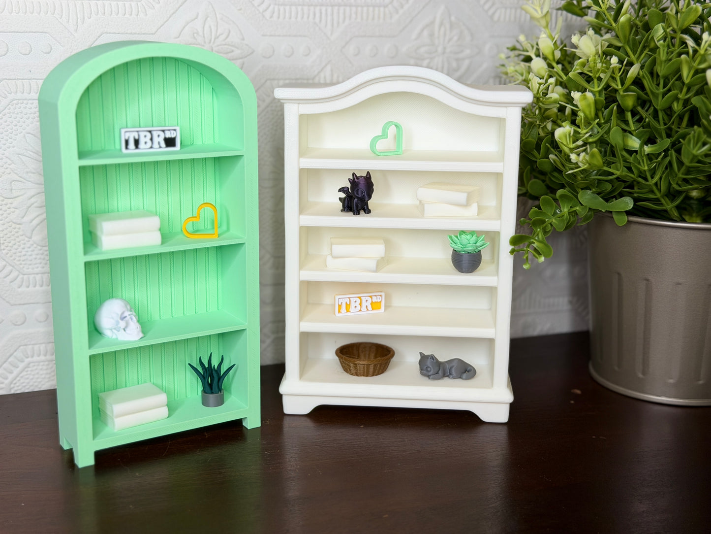 Two miniature decorative bookshelves, one mint green and one white, styled with tiny books, plants, and small figurines on a dark wooden surface against a textured white wall.
