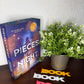The book 'Pieces of the Night' by Jennifer Hartmann displayed on a dark wooden table next to a potted green plant and colorful 'BOOK' letter cutouts.