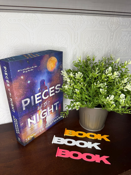 The book 'Pieces of the Night' by Jennifer Hartmann displayed on a dark wooden table next to a potted green plant and colorful 'BOOK' letter cutouts.