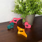 Four colorful plastic ring-shaped clips in pink, yellow, and teal scattered on a dark wooden table next to a potted green plant.