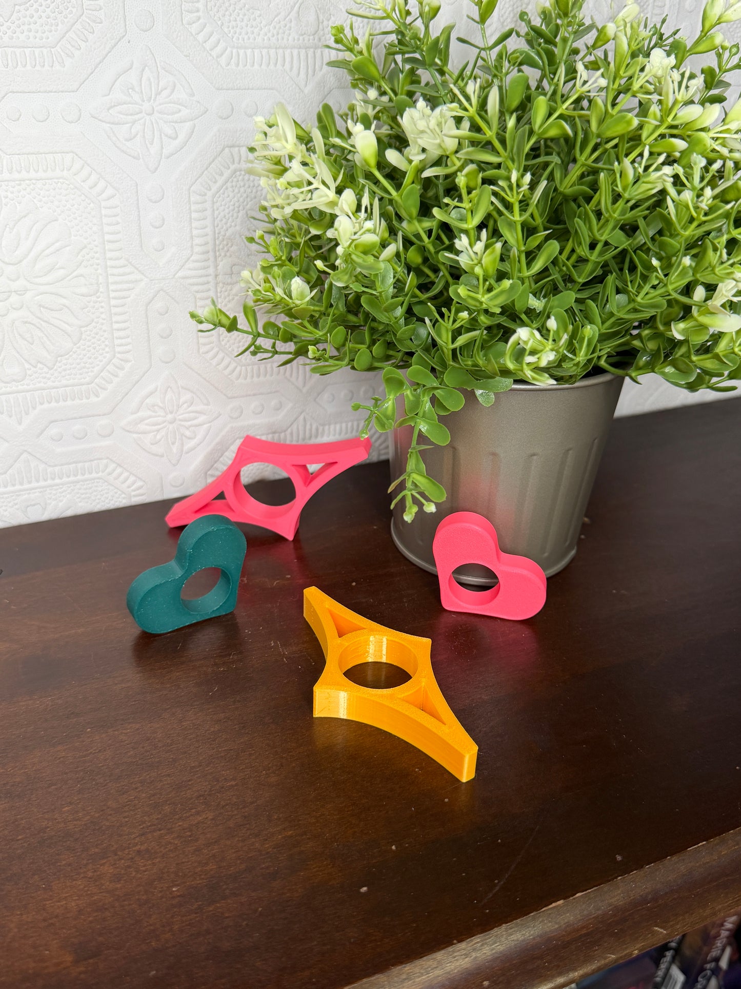 Four colorful plastic ring-shaped clips in pink, yellow, and teal scattered on a dark wooden table next to a potted green plant.