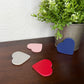 Four small heart-shaped items in blue, red, pink, and light gray are arranged on a dark wooden surface next to a white patterned surface and a potted green plant.