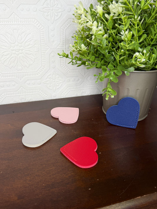 Four small heart-shaped items in blue, red, pink, and light gray are arranged on a dark wooden surface next to a white patterned surface and a potted green plant.