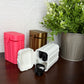 Three small hard plastic storage cases in white, metallic gold, and bright pink, arranged on a dark brown tabletop next to a potted green plant.