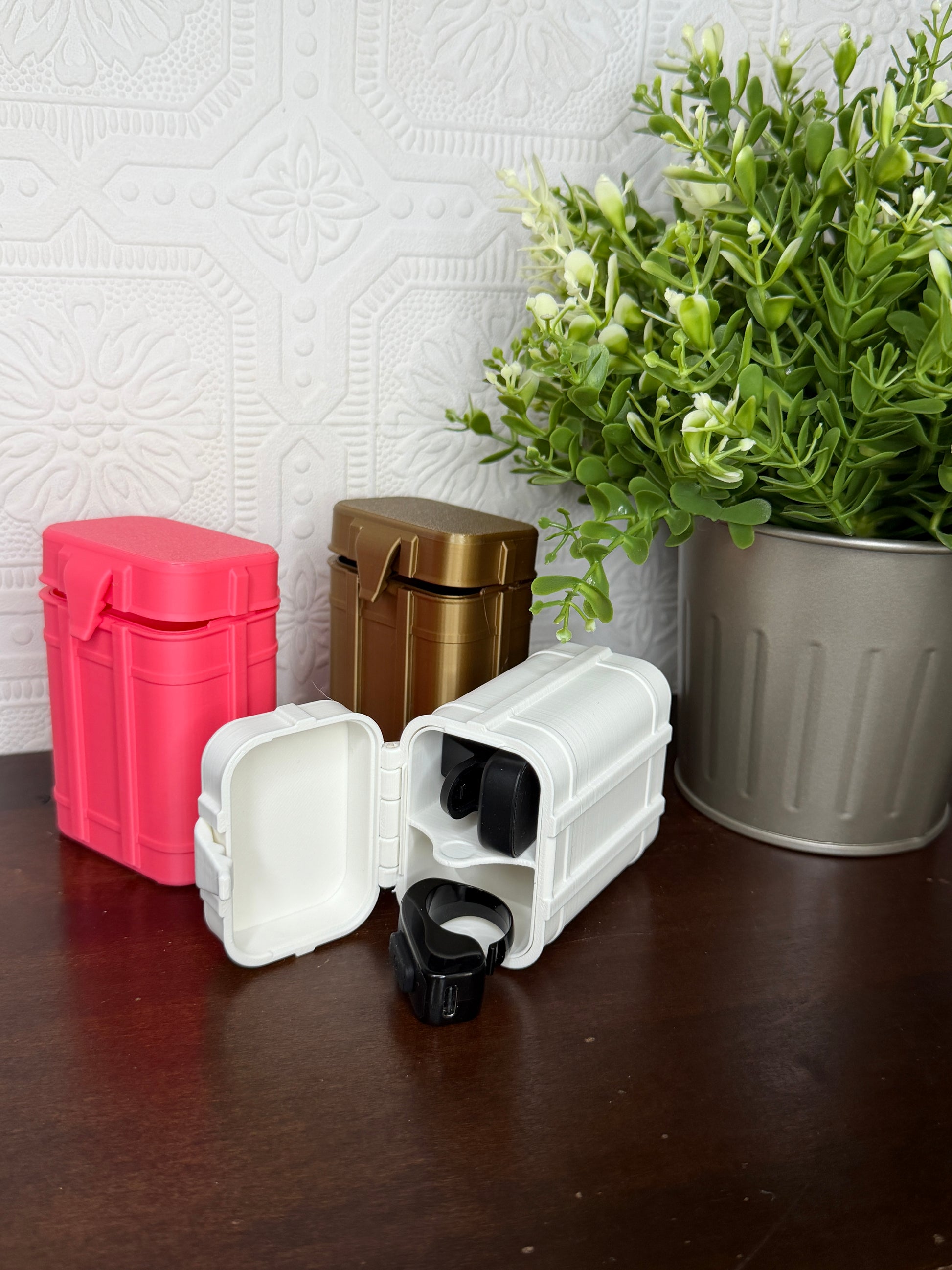 Three small hard plastic storage cases in white, metallic gold, and bright pink, arranged on a dark brown tabletop next to a potted green plant.