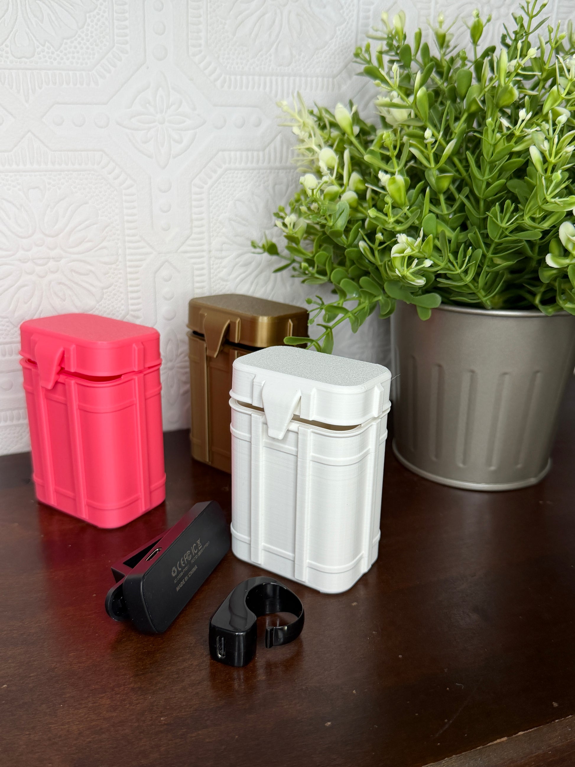 Three rectangular metal pill organizer boxes in pink, gold, and white, displayed on a dark wooden table next to a potted green plant. A black clip and a black ring clip are shown in front of the boxes.