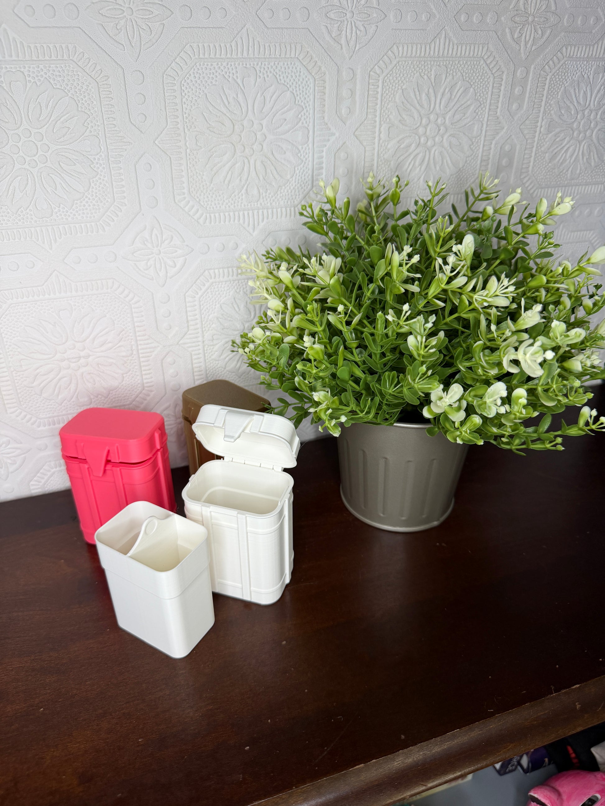 Three small plastic storage containers in white, pink, and brown, displayed on a dark wooden table next to a potted green plant against a textured light gray wall.