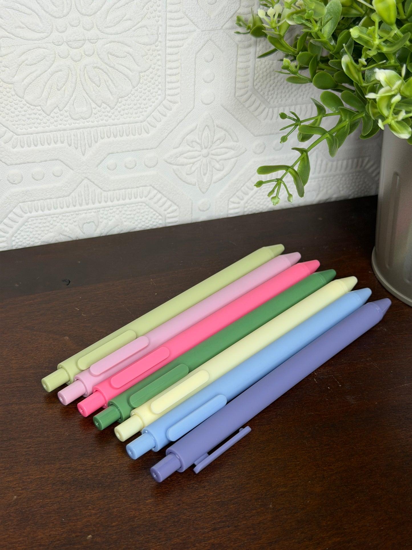 Seven pastel-colored retractable gel pens in shades of green, pink, yellow, blue, and purple, arranged diagonally on a dark wooden tabletop next to a potted green plant.